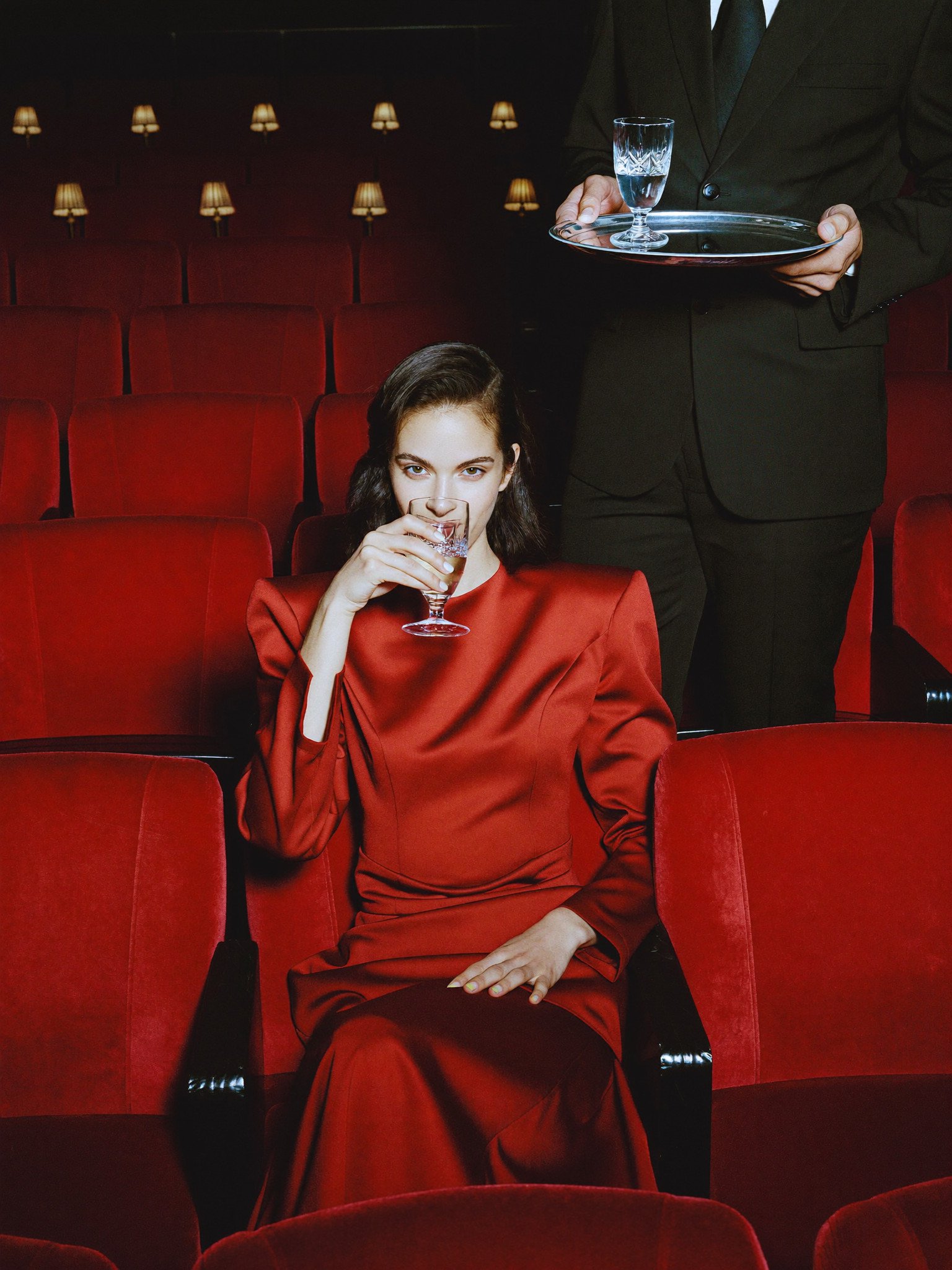 Woman in red drinking in a red velvet cinema, a waiter behind her