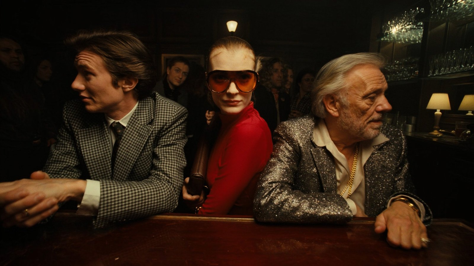 Three people at a dim bar, a woman in red sunglasses between two men